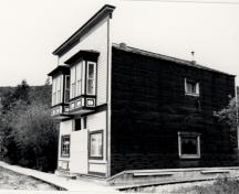 Vue de la façade générale de Ruby's Place, qui montre le parement de planches horizontales à gorge, peintes, de la façade sur rue, avec ses grands oriels caractéristiques, 1987.; Parks Canada Agency / Agence Parcs Canada, 1987.