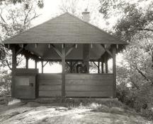 General view of the Adelaide Island Picnic Shelter, showing the hipped roof, exposed rafters, vertical wood support posts and wood brackets, 1992.; Parks Canada Agency / Agence Parcs Canada / Historica Resources Ltd., 1992.