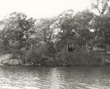 View of the Adelaide Island Picnic Shelter from the river, showing its relatively isolated island location on a heavily treed site, 1992.; Parks Canada Agency / Agence Parcs Canada / Historica Resources Ltd., 1992.